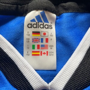 May include: A blue Adidas shirt with a white and black collar. The size tag shows the size in different countries, including D 152, J 150, GB LB, F, 1 12, USA M, E 152, CDN M/M.