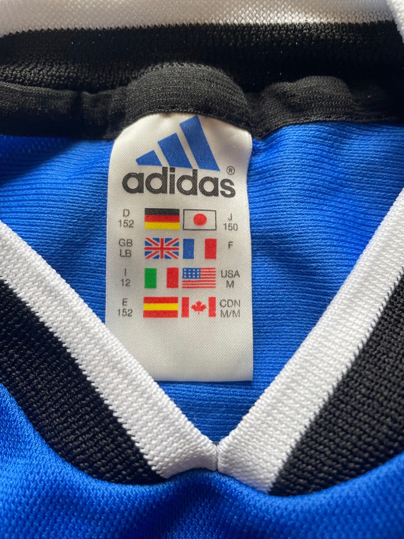 May include: A blue Adidas shirt with a white and black collar. The size tag shows the size in different countries, including D 152, J 150, GB LB, F, 1 12, USA M, E 152, CDN M/M.