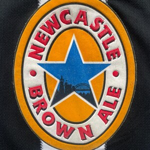May include: Close-up of a Newcastle Brown Ale patch. The patch is circular with a white border, an orange center, and a blue star. The words "NEWCASTLE BROWN ALE" are in red. The background is a black and white striped fabric.