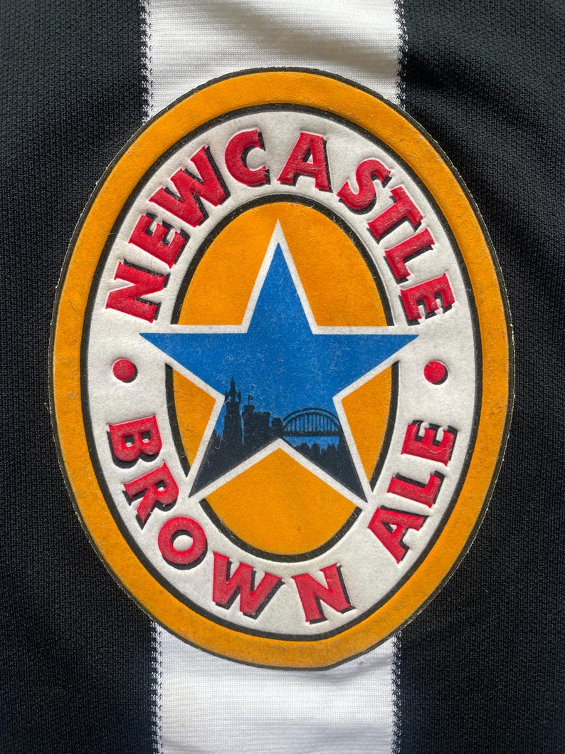 May include: Close-up of a Newcastle Brown Ale patch. The patch is circular with a white border, an orange center, and a blue star. The words "NEWCASTLE BROWN ALE" are in red. The background is a black and white striped fabric.
