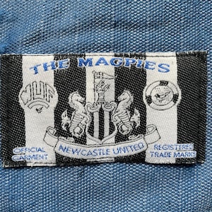May include: A black and white striped patch with the words "THE MAGPIES" in blue. The patch displays the Newcastle United crest, the text "NEWCASTLE UNITED", and the phrases "OFFICIAL GARMENT" and "REGISTERED TRADE MARKS".
