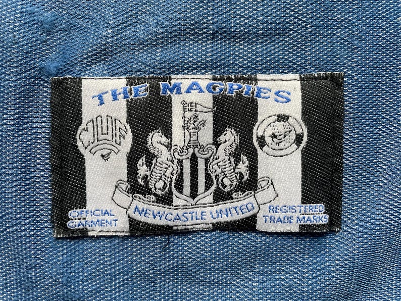May include: A black and white striped patch with the words "THE MAGPIES" in blue. The patch displays the Newcastle United crest, the text "NEWCASTLE UNITED", and the phrases "OFFICIAL GARMENT" and "REGISTERED TRADE MARKS".