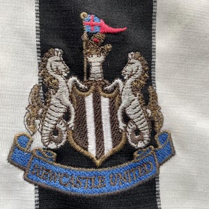 Puede incluir: Camiseta de f&uacute;tbol negra y blanca bordada con un escudo que presenta un escudo con tres rayas verticales, dos caballitos de mar y una bandera con una cruz. El texto "NEWCASTLE UNITED" est&aacute; en una cinta azul.