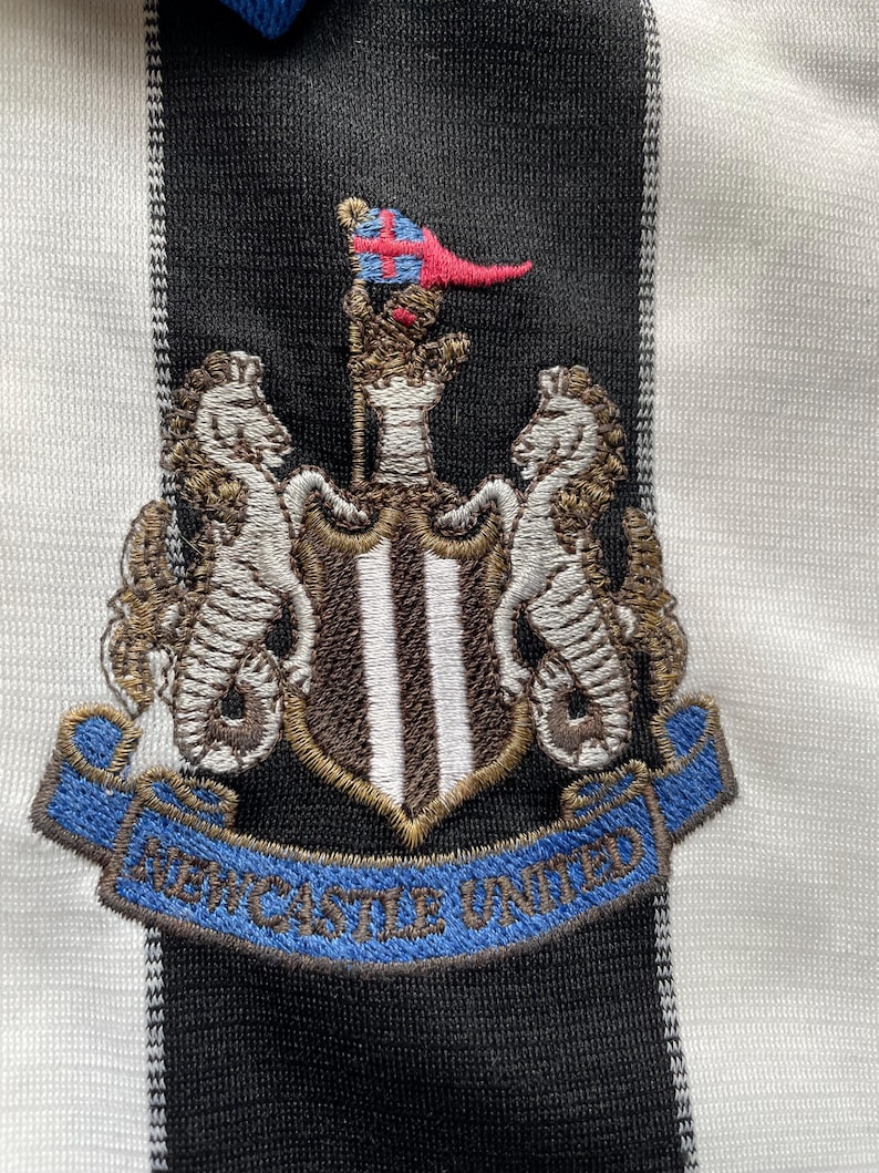 Puede incluir: Camiseta de f&uacute;tbol negra y blanca bordada con un escudo que presenta un escudo con tres rayas verticales, dos caballitos de mar y una bandera con una cruz. El texto "NEWCASTLE UNITED" est&aacute; en una cinta azul.