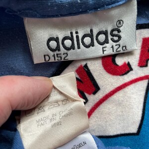 May include: A blue adidas t-shirt with a white and red logo that says "N.C.A." The tag says "adidas" with the size "D152" and "F 120". The tag also says "Made in China" and "Fait en Chine" with the number "9692". The tag also says "Waschen und B&uuml;geln von Links Bunt: Bitte Feinwaschmittel".