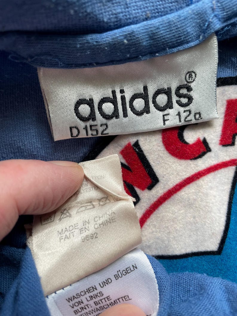 May include: A blue adidas t-shirt with a white and red logo that says "N.C.A." The tag says "adidas" with the size "D152" and "F 120". The tag also says "Made in China" and "Fait en Chine" with the number "9692". The tag also says "Waschen und B&uuml;geln von Links Bunt: Bitte Feinwaschmittel".