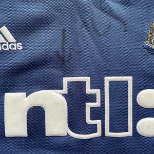 Op de afbeelding: Een blauw voetbaltenue met een wit Adidas-logo, een wit "ntl"-logo en een zwart-wit embleem van Newcastle United Football Club.