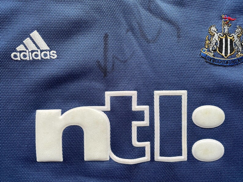 Op de afbeelding: Een blauw voetbaltenue met een wit Adidas-logo, een wit "ntl"-logo en een zwart-wit embleem van Newcastle United Football Club.