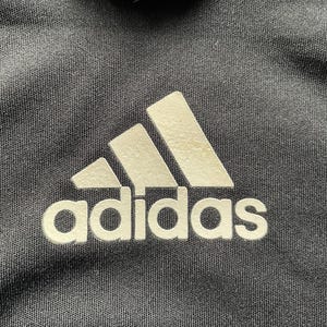 Pu&ograve; includere: Primo piano di una maglietta sportiva nera con il logo Adidas in bianco. Il logo presenta tre strisce bianche sopra la parola "adidas". La maglietta ha un colletto bianco.