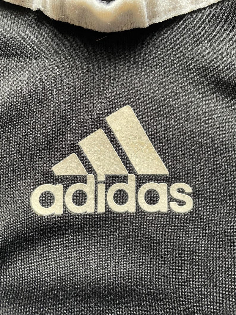 Pu&ograve; includere: Primo piano di una maglietta sportiva nera con il logo Adidas in bianco. Il logo presenta tre strisce bianche sopra la parola "adidas". La maglietta ha un colletto bianco.