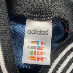 May include: Close-up of an Adidas tag. The tag is white with black text and features the Adidas logo. It includes size and country codes, with flags for Germany, Great Britain, the United States, and others. The background is blue.