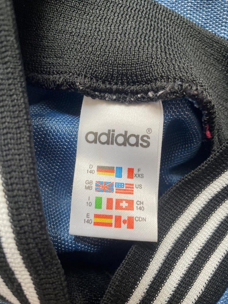 May include: Close-up of an Adidas tag. The tag is white with black text and features the Adidas logo. It includes size and country codes, with flags for Germany, Great Britain, the United States, and others. The background is blue.