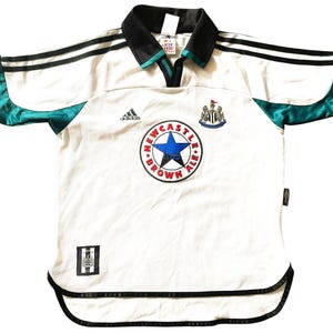 Pu&ograve; includere: Maglia da calcio bianca con dettagli neri e verdi. La maglia ha il logo Adidas, uno stemma del Newcastle United e il testo "NEWCASTLE BROWN ALE" in un cerchio.