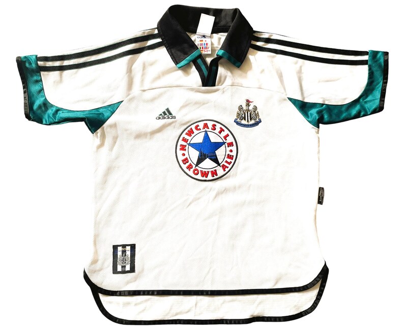 Pu&ograve; includere: Maglia da calcio bianca con dettagli neri e verdi. La maglia ha il logo Adidas, uno stemma del Newcastle United e il testo "NEWCASTLE BROWN ALE" in un cerchio.