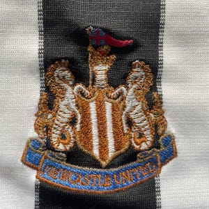 May include: Close-up of a black and white striped fabric with an embroidered crest. The crest features two seahorses flanking a shield with a banner that reads "NEWCASTLE UNITED" in blue lettering.