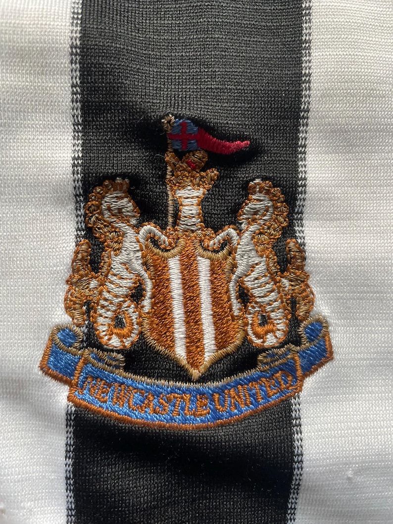 May include: Close-up of a black and white striped fabric with an embroidered crest. The crest features two seahorses flanking a shield with a banner that reads "NEWCASTLE UNITED" in blue lettering.