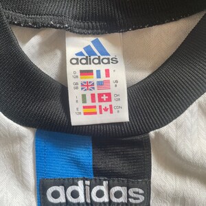 May include: White adidas football shirt with blue and black stripes. The shirt has a size tag with the adidas logo and size information in different languages and measurements. The tag reads: "D 128, GB SB, E 128, F, US 8, CH 128, CDN 8".