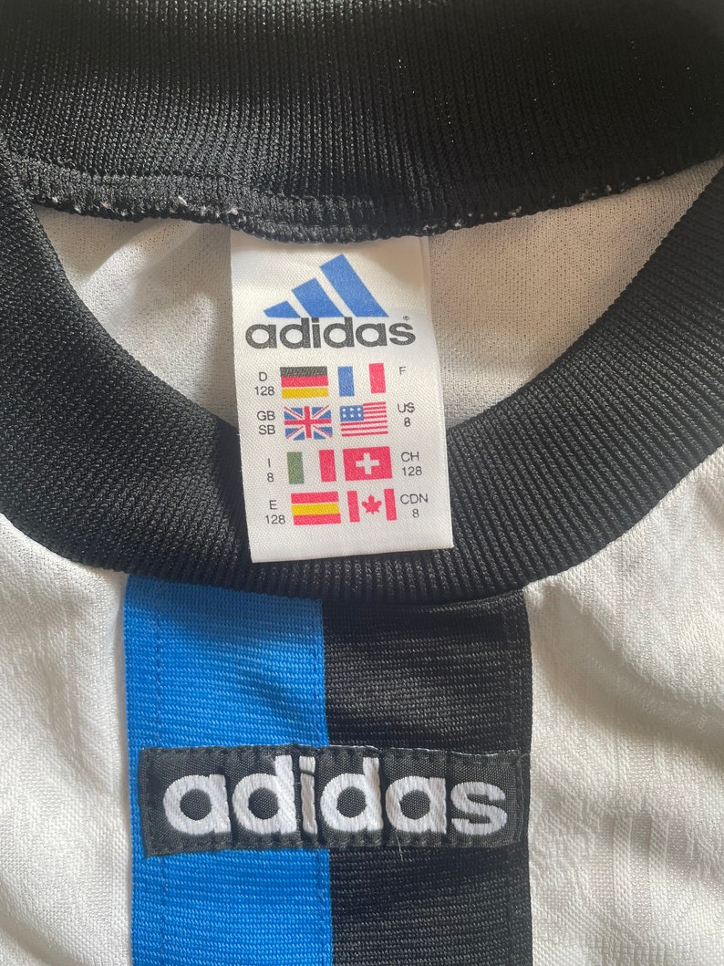 May include: White adidas football shirt with blue and black stripes. The shirt has a size tag with the adidas logo and size information in different languages and measurements. The tag reads: "D 128, GB SB, E 128, F, US 8, CH 128, CDN 8".