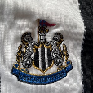 May include: A white and black striped football shirt with a black and white embroidered crest featuring a shield with black and white stripes, two gold seahorses, and a gold banner with the text "Newcastle United".