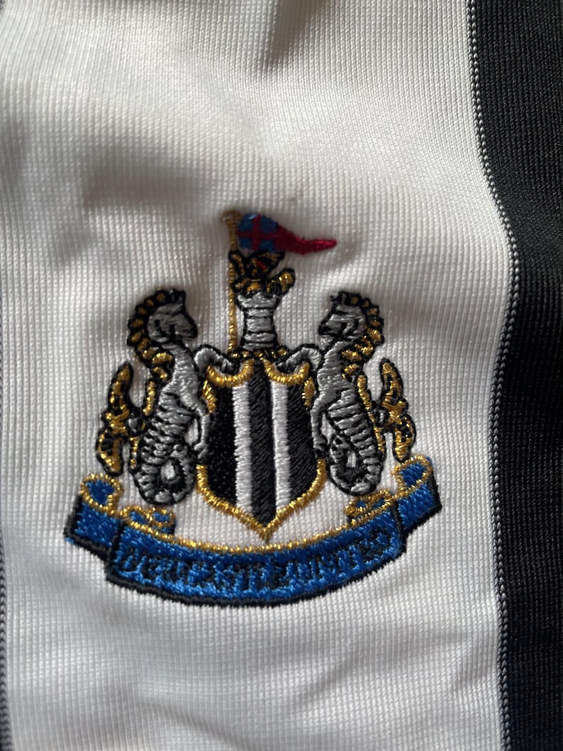 May include: A white and black striped football shirt with a black and white embroidered crest featuring a shield with black and white stripes, two gold seahorses, and a gold banner with the text "Newcastle United".
