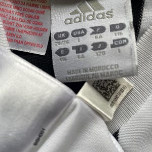 May include: Close-up of a white Adidas tag with black stripes. The tag includes the Adidas logo, size information, and text in multiple languages. The tag reads "Made in Morocco" and "Fabriqu&eacute; au Maroc".