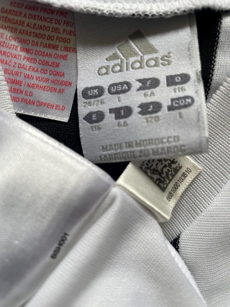 May include: Close-up of a white Adidas tag with black stripes. The tag includes the Adidas logo, size information, and text in multiple languages. The tag reads "Made in Morocco" and "Fabriqu&eacute; au Maroc".