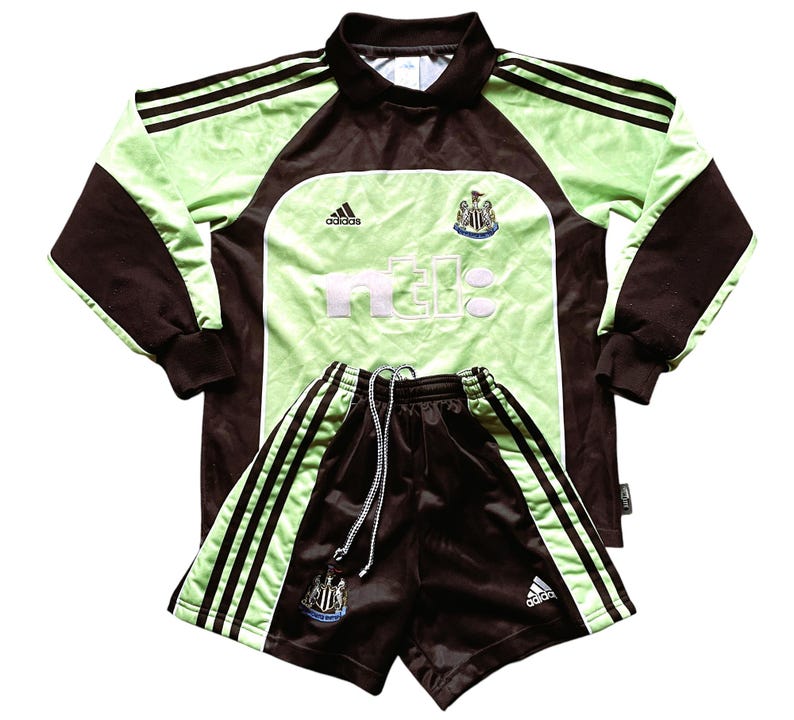 May include: A green and brown soccer jersey and shorts set with the Newcastle United logo and the word "nufc" on the front. The jersey is long-sleeved and has Adidas branding. The shorts have three white stripes on each side.