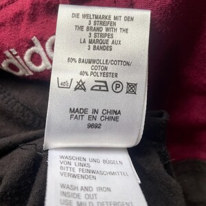 Puede incluir: Primer plano de una etiqueta de ropa con texto en varios idiomas. La etiqueta incluye el nombre de la marca y las instrucciones de lavado. La tela es de color burdeos oscuro con una tela negra debajo.