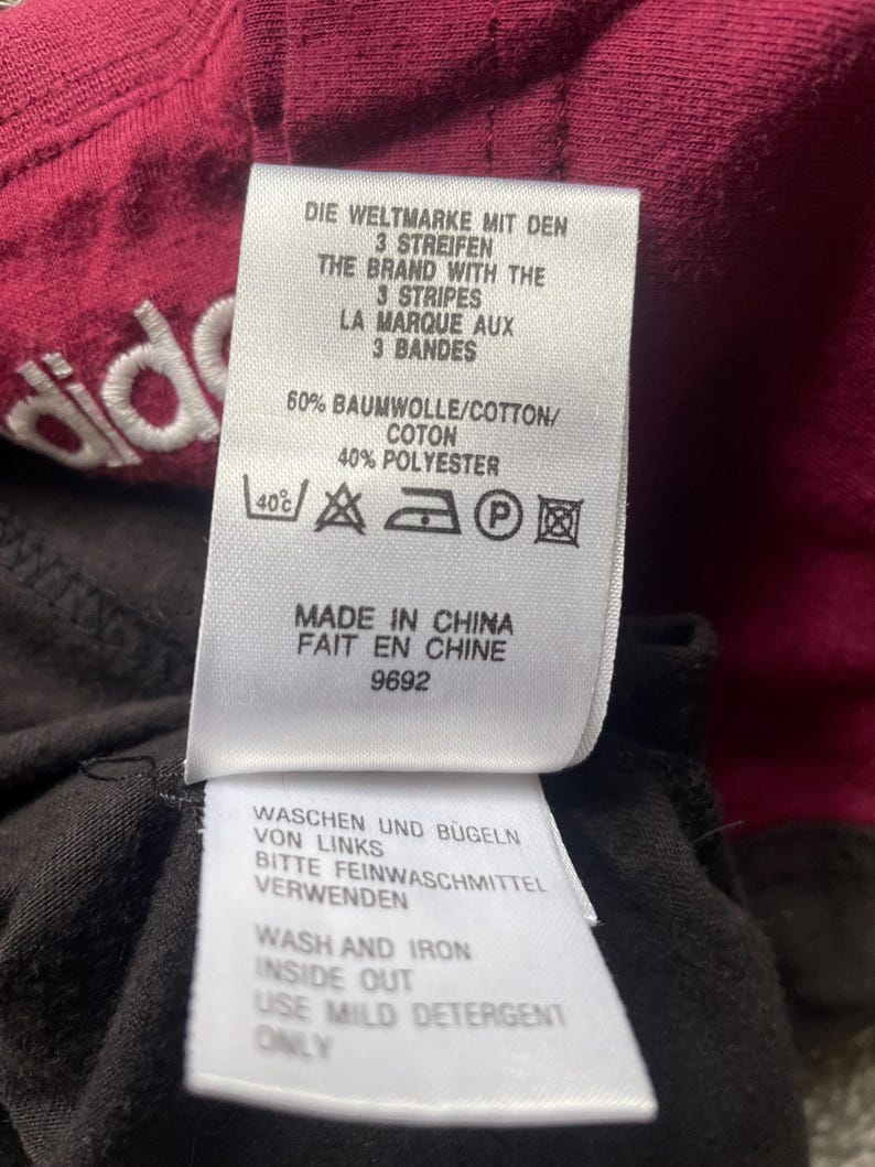 Puede incluir: Primer plano de una etiqueta de ropa con texto en varios idiomas. La etiqueta incluye el nombre de la marca y las instrucciones de lavado. La tela es de color burdeos oscuro con una tela negra debajo.