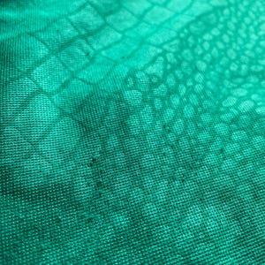 K&ouml;nnte beinhalten: Nahaufnahme eines strukturierten, smaragdgr&uuml;nen Stoffes mit einem Reptilienschuppenmuster. Der Stoff hat ein leicht abgenutztes Aussehen mit subtilen Farb- und Texturvariationen. Das Material scheint ein Strick- oder Webtextil zu sein.