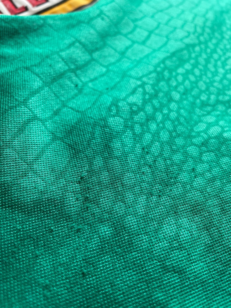 K&ouml;nnte beinhalten: Nahaufnahme eines strukturierten, smaragdgr&uuml;nen Stoffes mit einem Reptilienschuppenmuster. Der Stoff hat ein leicht abgenutztes Aussehen mit subtilen Farb- und Texturvariationen. Das Material scheint ein Strick- oder Webtextil zu sein.