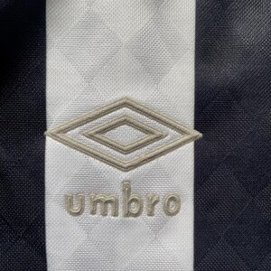 Op de afbeelding: Close-up van een zwart-wit sportshirt. Een witte verticale streep loopt door het midden, met het Umbro-logo en de tekst in lichtbruin. Het shirt heeft een textuurpatroon.