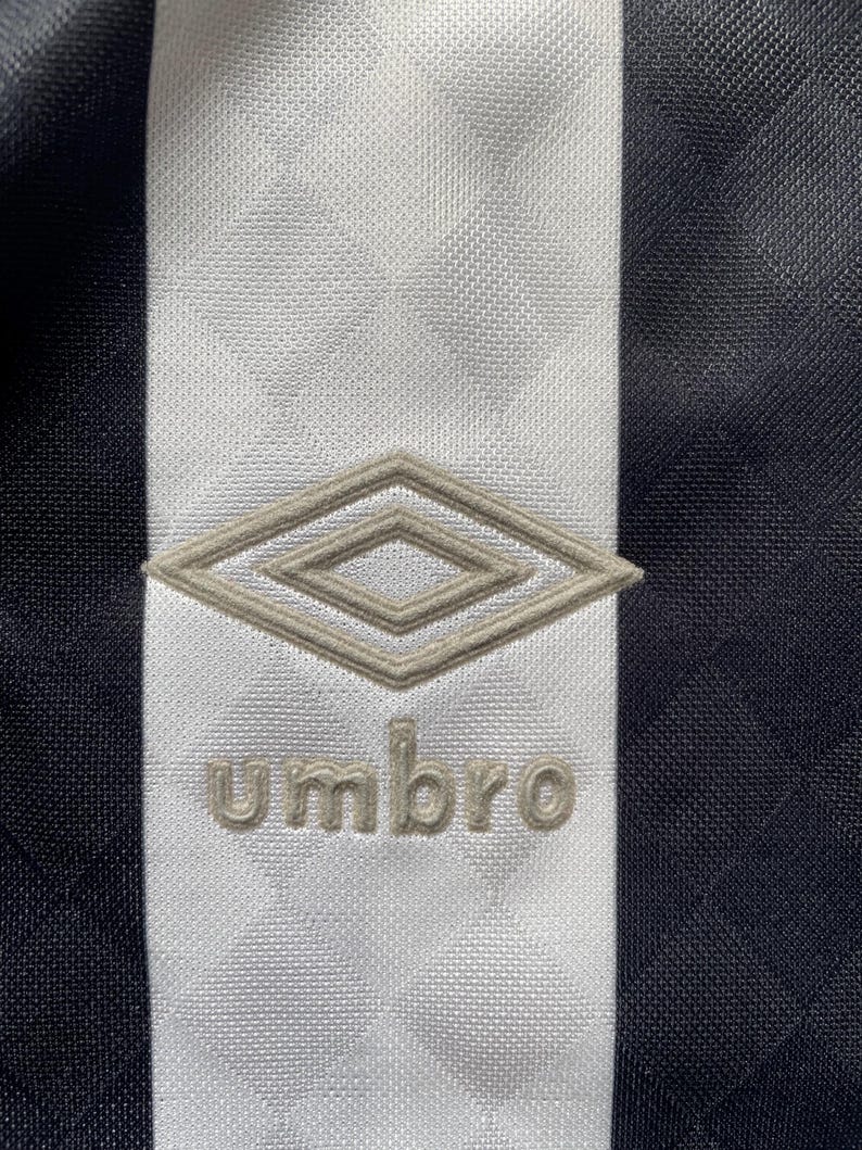 Op de afbeelding: Close-up van een zwart-wit sportshirt. Een witte verticale streep loopt door het midden, met het Umbro-logo en de tekst in lichtbruin. Het shirt heeft een textuurpatroon.