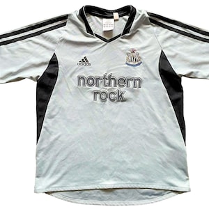 Pu&ograve; includere: Una maglia da calcio blu chiaro con dettagli neri. La maglia ha il logo Adidas sul lato destro e lo stemma del Newcastle United sul lato sinistro. Il testo "northern rock" &egrave; stampato in grigio sul davanti della maglia.