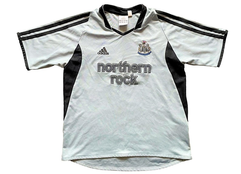 Pu&ograve; includere: Una maglia da calcio blu chiaro con dettagli neri. La maglia ha il logo Adidas sul lato destro e lo stemma del Newcastle United sul lato sinistro. Il testo "northern rock" &egrave; stampato in grigio sul davanti della maglia.