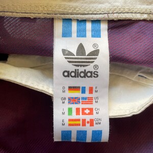 May include: Close-up of an Adidas tag with the brand's logo and text. The tag features the Adidas logo, a trefoil, and the brand name. The tag also includes flags representing different countries, including the United States, Great Britain, and Canada.