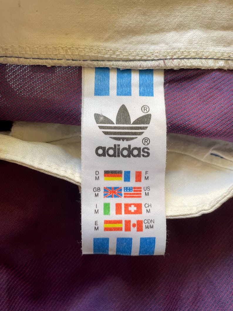 May include: Close-up of an Adidas tag with the brand's logo and text. The tag features the Adidas logo, a trefoil, and the brand name. The tag also includes flags representing different countries, including the United States, Great Britain, and Canada.