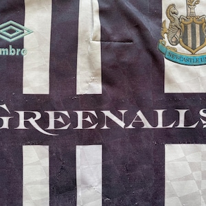 Pode incluir: Camisola de futebol de riscas pretas e brancas com o bras&atilde;o do Newcastle United e a palavra "GREENALL'S" impressa na frente. A camisola tamb&eacute;m apresenta o logotipo da Umbro.