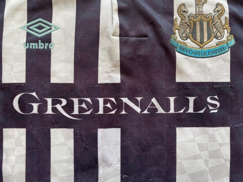 Pode incluir: Camisola de futebol de riscas pretas e brancas com o bras&atilde;o do Newcastle United e a palavra "GREENALL'S" impressa na frente. A camisola tamb&eacute;m apresenta o logotipo da Umbro.