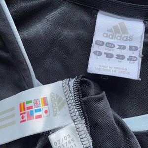 May include: Black Adidas track jacket with a white zipper and white piping. The jacket has a tag with the Adidas logo and the text "MADE IN TUNISIA FABRIQUE EN TUNISIE".