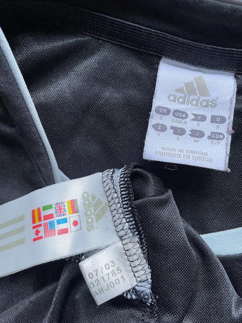 May include: Black Adidas track jacket with a white zipper and white piping. The jacket has a tag with the Adidas logo and the text "MADE IN TUNISIA FABRIQUE EN TUNISIE".