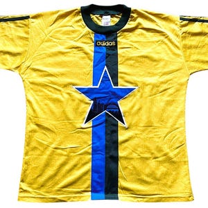 May include: A yellow soccer jersey with a blue and black star design on the front. The jersey has a blue stripe down the center and the Adidas logo at the top. The sleeves have black stripes.