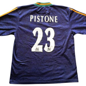 May include: A navy blue soccer jersey with the name "Pistone" and the number "23" in white. The jersey has green and orange accents on the collar and sleeves. The fabric has a subtle pattern.