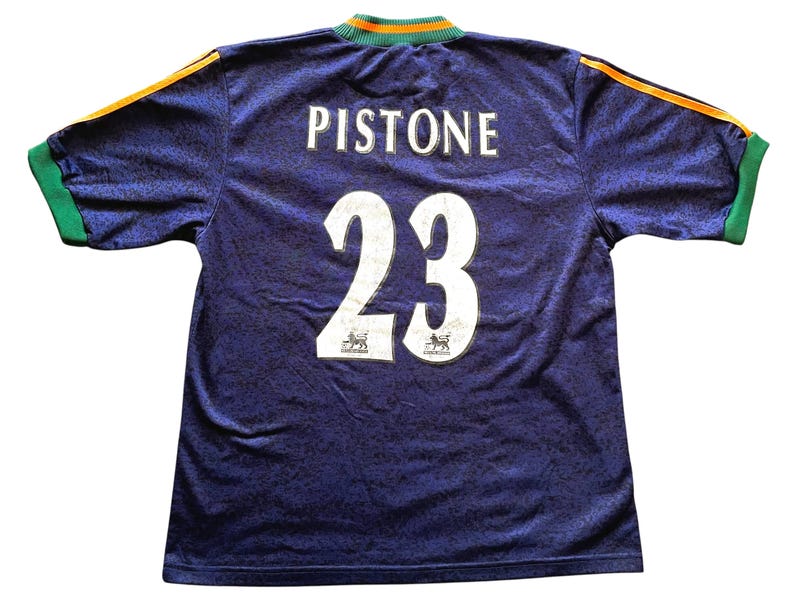 May include: A navy blue soccer jersey with the name "Pistone" and the number "23" in white. The jersey has green and orange accents on the collar and sleeves. The fabric has a subtle pattern.
