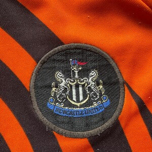 May include: A black and white embroidered patch with the Newcastle United crest. The crest features a shield with two black and white seahorses on either side. The shield is topped with a flag and a crown. The words "Newcastle United" are written in a blue ribbon below the shield.
