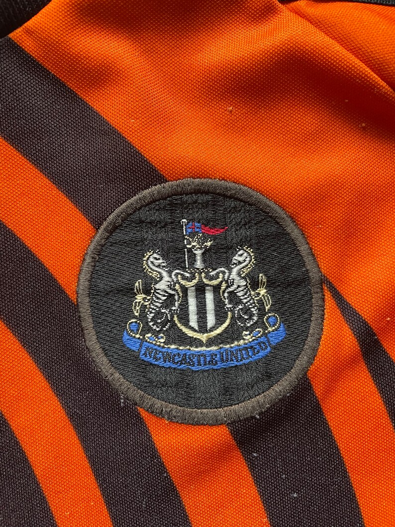 May include: A black and white embroidered patch with the Newcastle United crest. The crest features a shield with two black and white seahorses on either side. The shield is topped with a flag and a crown. The words "Newcastle United" are written in a blue ribbon below the shield.