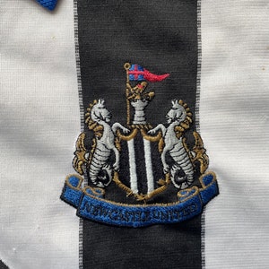 Może przedstawiać: Czarno-biała koszulka piłkarska w czarno-białe pasy. Koszulka ma niebieski kołnierzyk i haftowany czarno-biały herb z napisem "NEWCASTLE UNITED".
