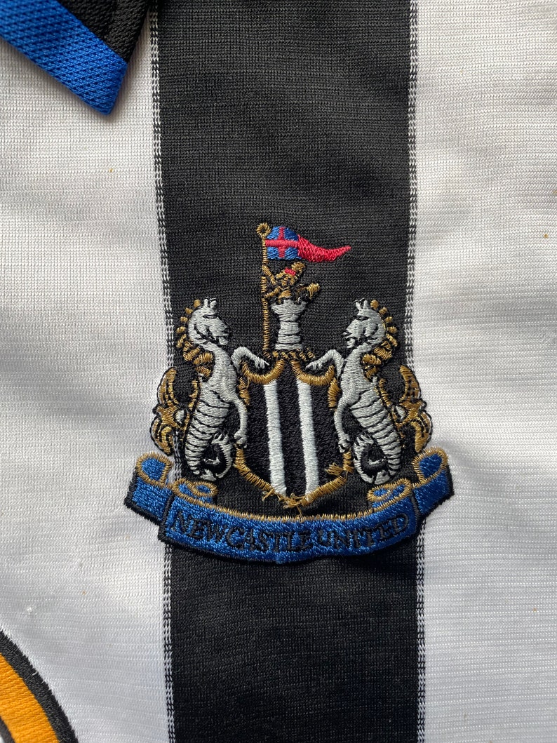 Może przedstawiać: Czarno-biała koszulka piłkarska w czarno-białe pasy. Koszulka ma niebieski kołnierzyk i haftowany czarno-biały herb z napisem "NEWCASTLE UNITED".