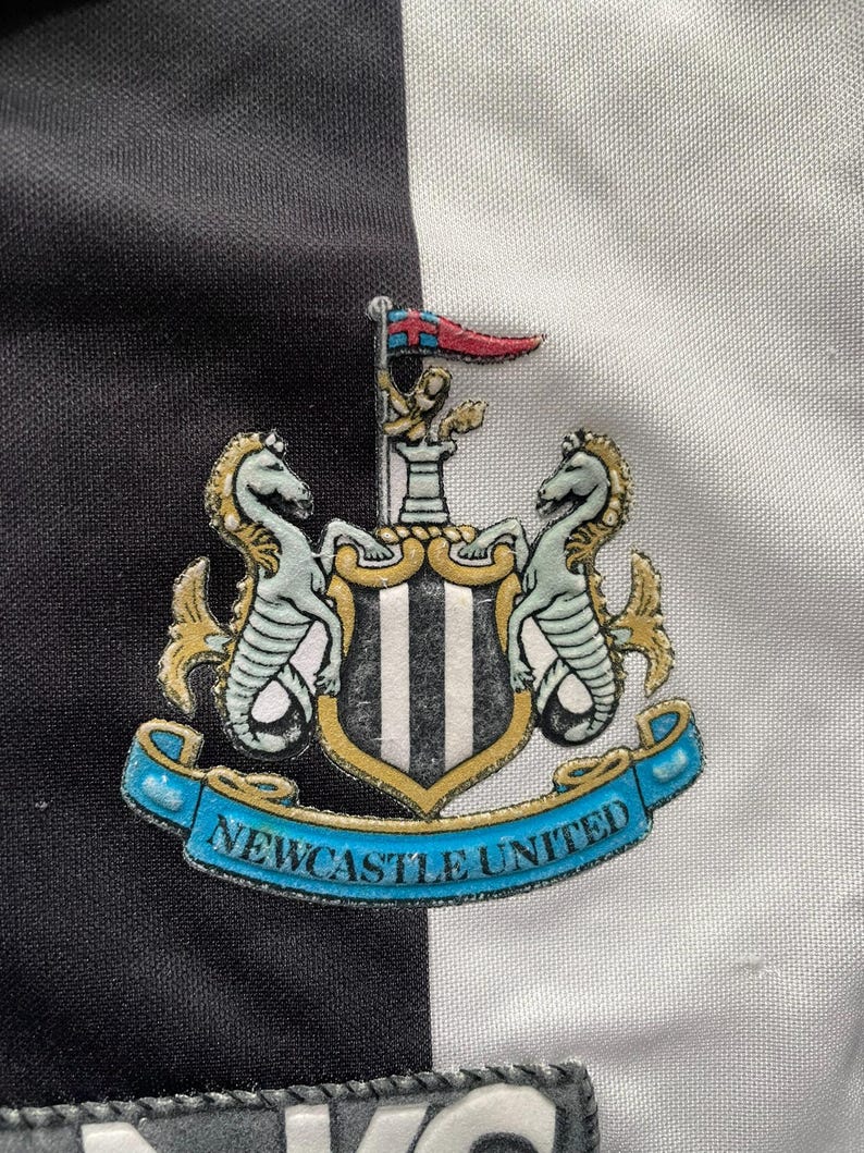 K&ouml;nnte beinhalten: Nahaufnahme eines Newcastle United-Wappens auf einem schwarz-wei&szlig; gestreiften Hintergrund. Das Wappen zeigt zwei Seepferdchen, einen Schild mit vertikalen Streifen und ein Banner mit der Aufschrift "Newcastle United".
