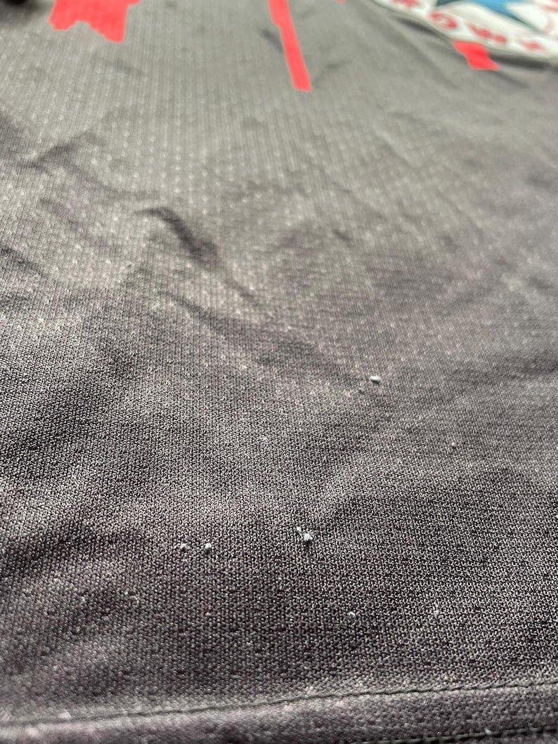 May include: Close-up of a black athletic shirt with a red design. The fabric has a textured, dotted pattern. The shirt appears to be made of a breathable material, suitable for sports or workouts.